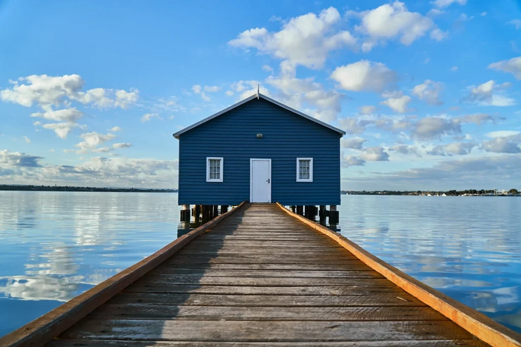 Blue boathouse at the end of a pier over a lake with a blue cloudy sky.