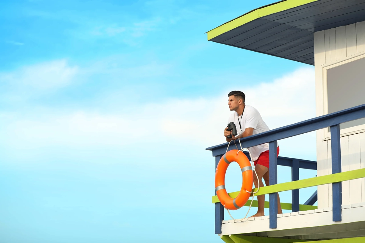 Male lifeguard on a tower looking out and holding binoculars.